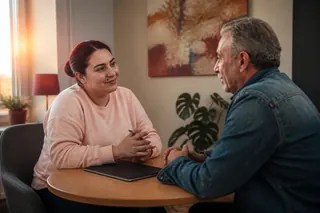 Pembe sweatshirtlü, topuz saçlı bir psikiyatrist ile bıyıklı, kot ceketli yaşlı bir adam öğleden sonra ışığında küçük yuvarlak bir masada sıcak bir sohbet ediyor